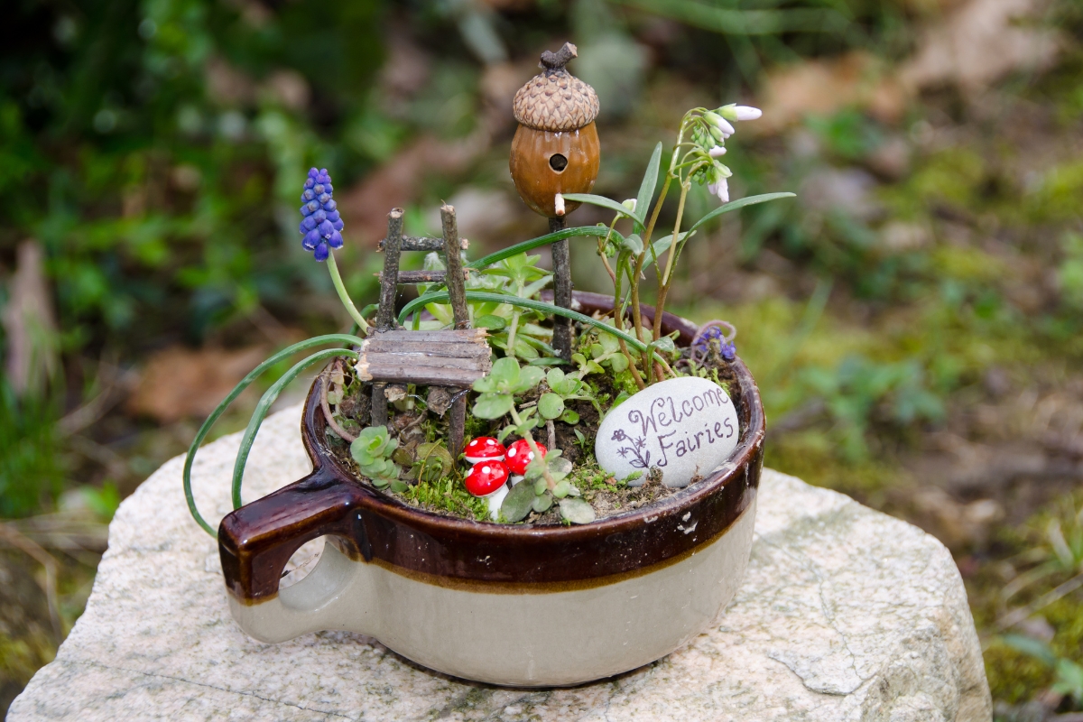 A fairy garden inside a shallow stoneware pot with small plants, succulents and furniture made from sticks. A stone that say's "Welcome Fairies" is placed to the right alongisde mushroom and acorn figures.