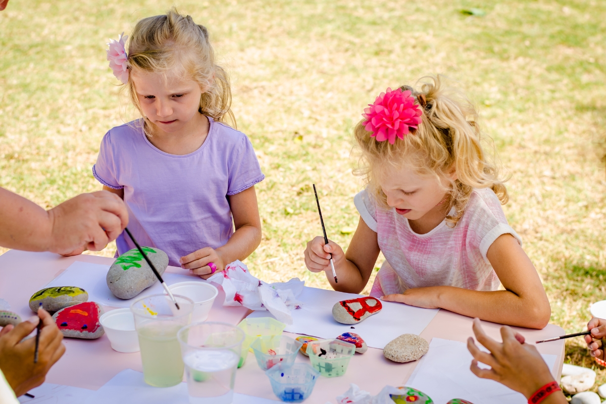 Two children sitting outside at a table painting on rocks.